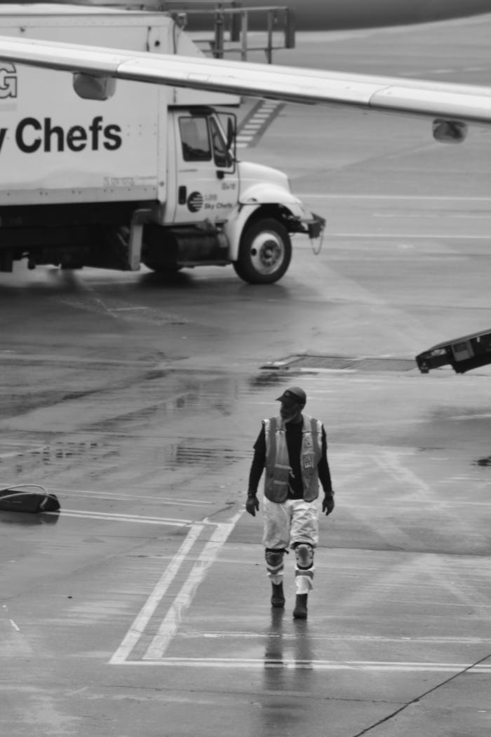 A lone airport worker in safety gear walking on a rain-soaked tarmac near a catering truck.